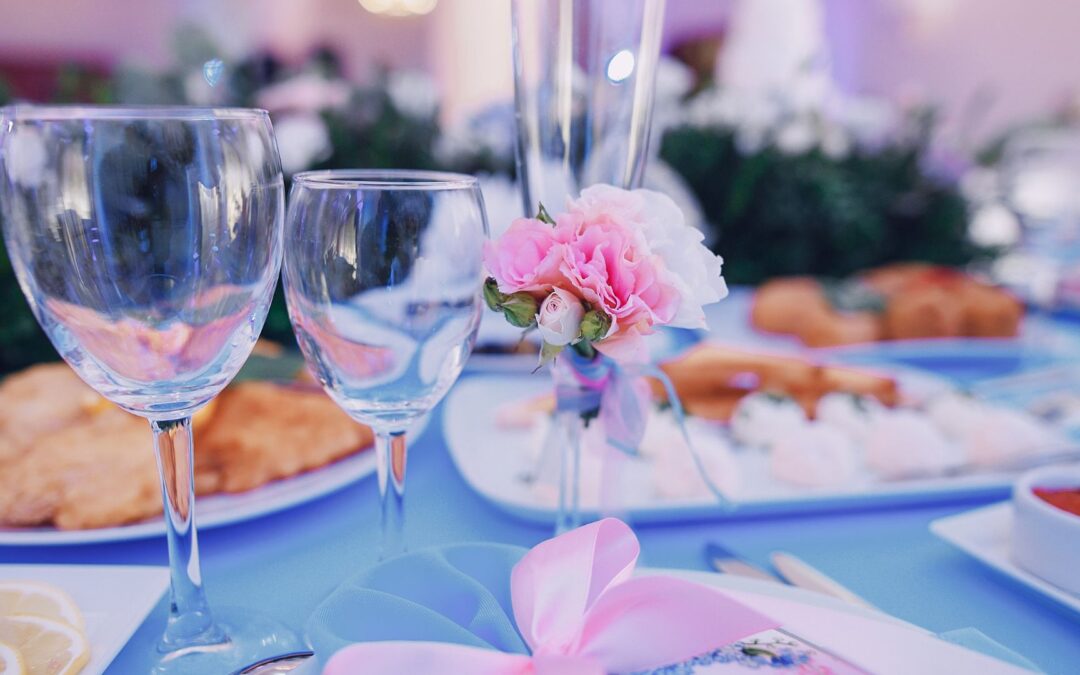 a pair of wine glasses and a pink carnation sit at the center of a table, with dishes piled high with food in the background