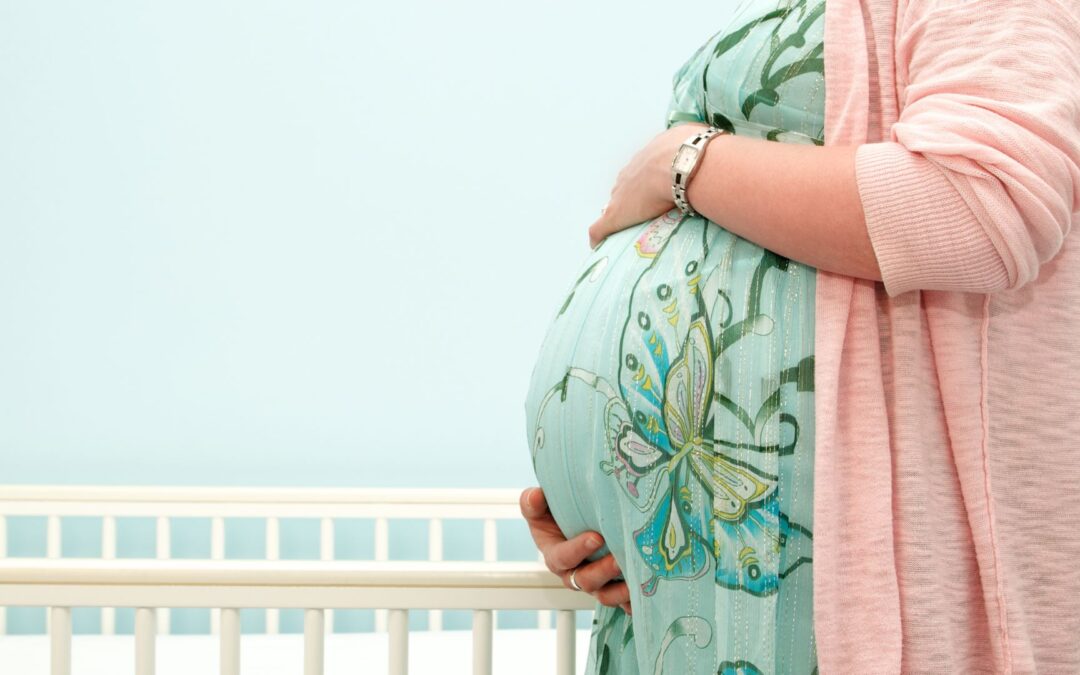a woman holds her very pregnant belly as she stands next to a crib in a lightly colored room