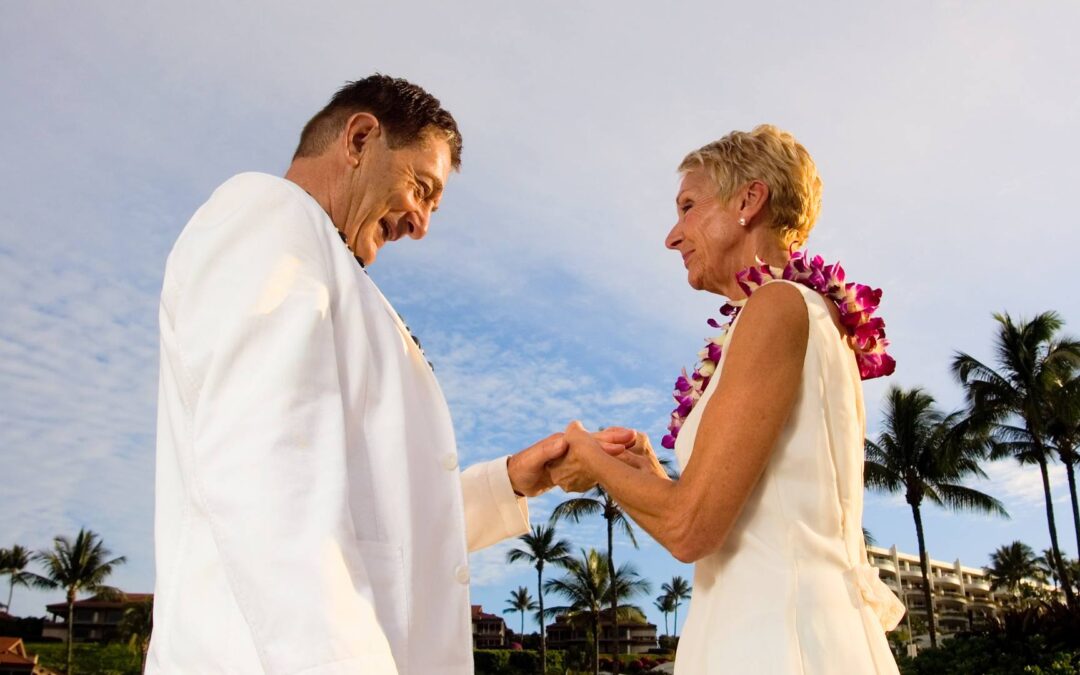 An older couple stand on the beach and look lovingly at each other, demonstrating a vow renewal.